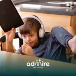 Autistic boy wearing headphones & holding a tablet at a kitchen table, illustrating screen use & modern childhood activities.