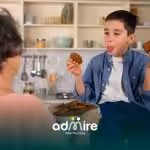 Autistic boy holding cookies & sticking out his tongue playfully while an adult offers a plate of baked treats in the kitchen.