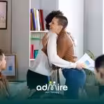 Two therapists hug in a modern office while teammates applaud, celebrating a successful project presentation.