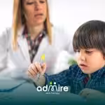 Autistic boy focusing on drawing shapes with a marker while a therapist or teacher guides him during a learning session.