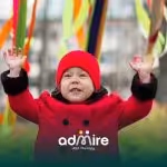 Toddler in a red coat and hat raising both hands outdoors with colorful ribbons hanging in the background.