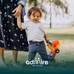 Autistic toddler holding a parent’s hand while standing on grass outdoors, holding a colorful toy truck.