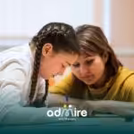 Autistic girl with braids focused on writing in a notebook while a supportive therapist watches over, helping her learn.