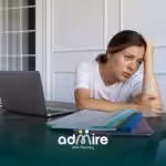 Stressed ABA women sitting at a desk with a laptop and folders, holding her head while worrying about responsibilities.