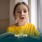Autistic child in a yellow shirt reading a book by a window and speaking aloud, practicing reading and language skills.