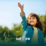 Happy autistic child outdoors raising one hand excitedly while smiling in a sunny field with green trees in the background.