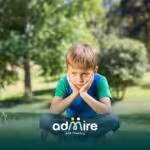 Autistic boy sitting alone on grass in a park with hands on his face, appearing sad or frustrated during a quiet moment.