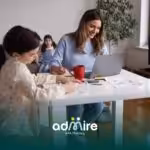 Therapist working on laptop at home while autistic girl draw at table, representing home-based ABA therapy.