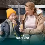 A therapist & an autistic boy sit together outdoors, sharing a snack, smiling and bonding during a relaxed daytime outing