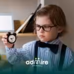 An autistic boy wearing glasses & suspenders carefully examines an alarm clock indoors, focusing on time awareness & learning