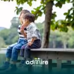 Two autistic children sitting on a wooden dock by a lake, sharing a frozen treat and enjoying time together outdoors.