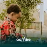 Autistic boy sitting outdoors on grass, focused on an object in hand while enjoying independent play in a garden setting.
