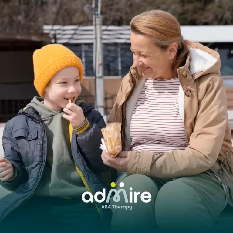 A therapist & an autistic boy sit together outdoors, sharing a snack, smiling and bonding during a relaxed daytime outing