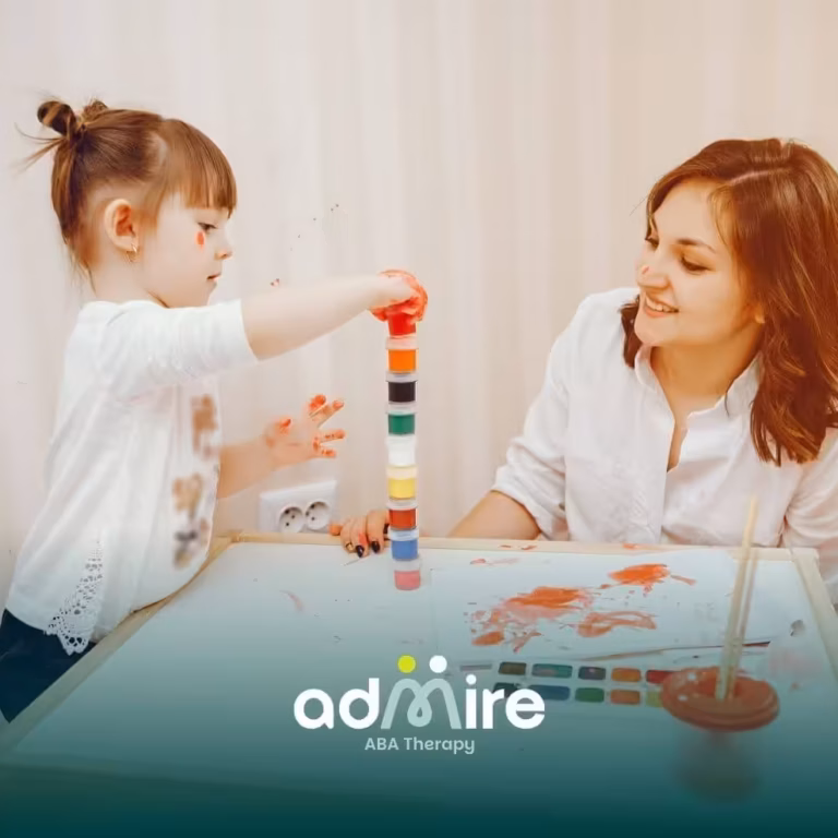 An autistic girl places a red cap on top of a colorful stack while her therapist watches with a warm smile