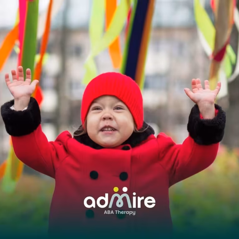 Toddler in a red coat and hat raising both hands outdoors with colorful ribbons hanging in the background.