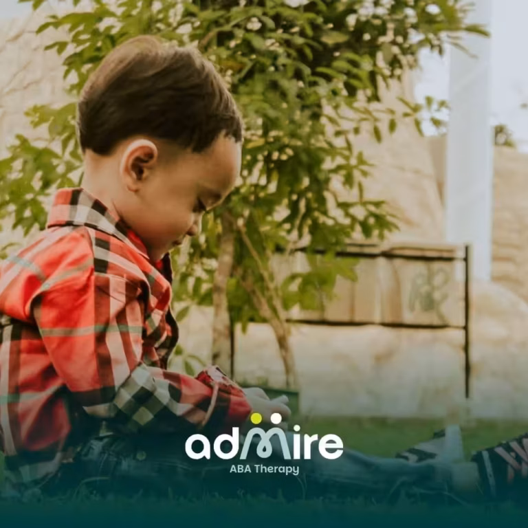 Autistic boy sitting outdoors on grass, focused on an object in hand while enjoying independent play in a garden setting.